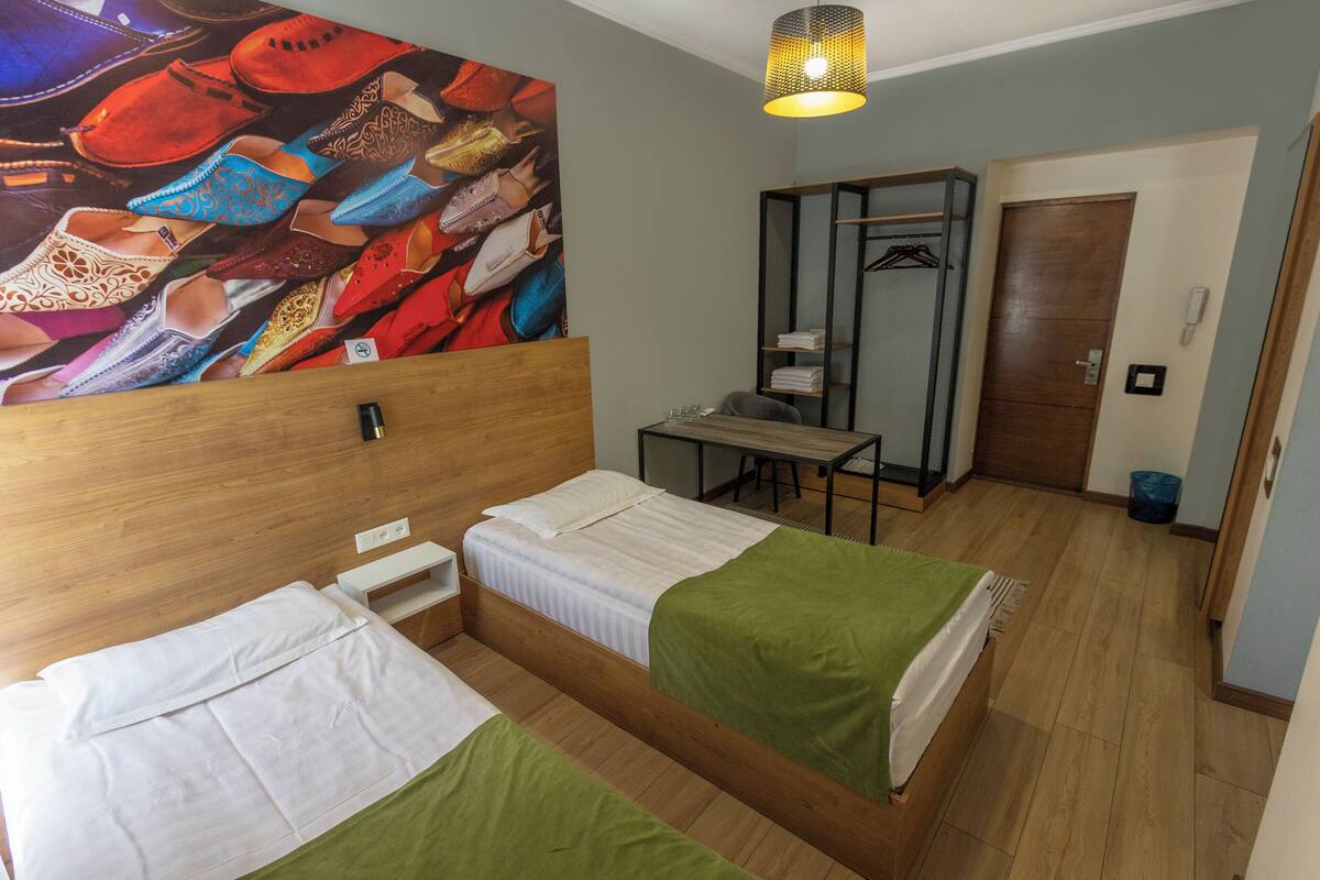 A hotel room with two beds, a desk, a chair, a wardrobe, and a large wall art piece.