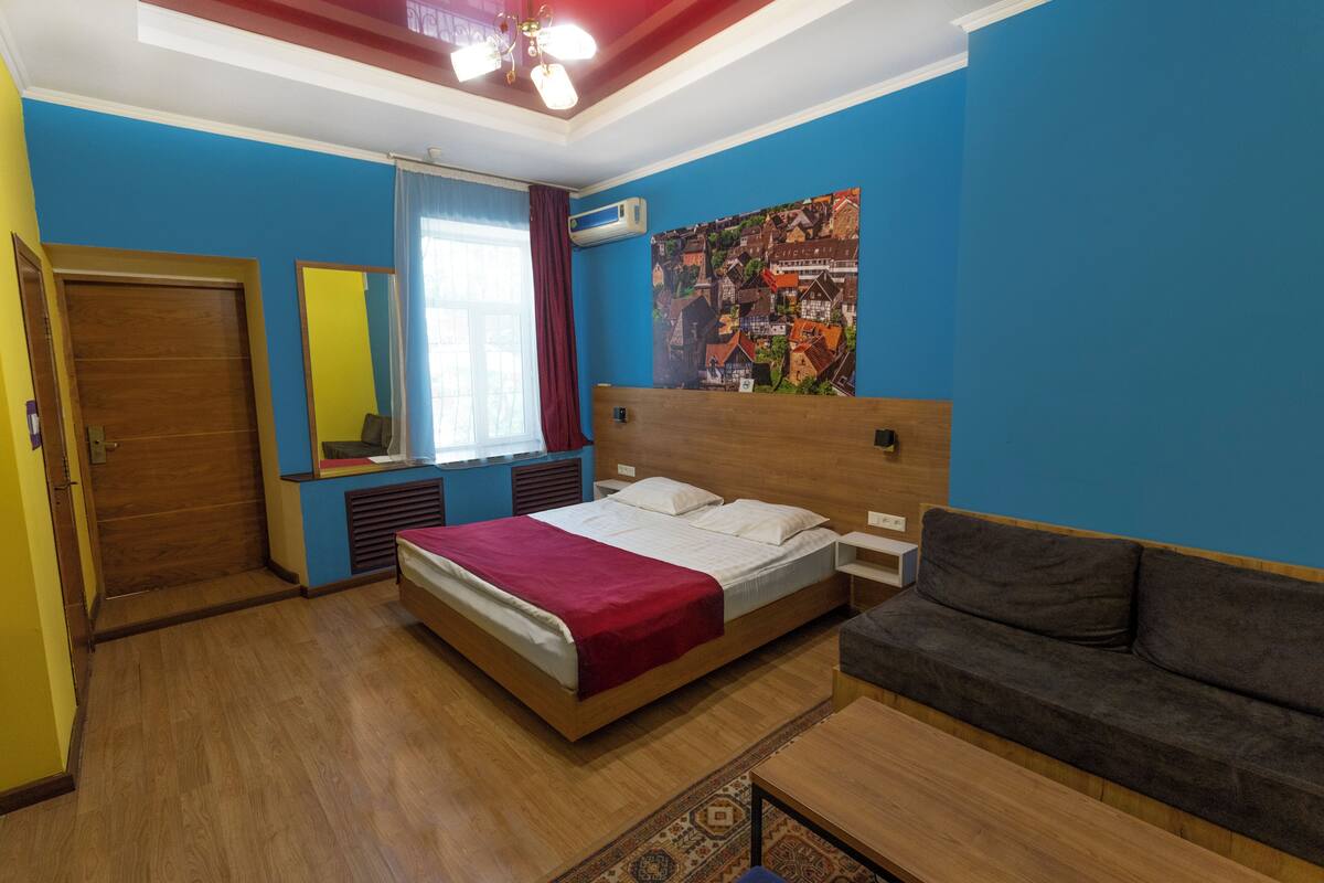 A bedroom with a wooden bed, a gray sofa, a large window with curtains, and a wall-mounted air conditioner.