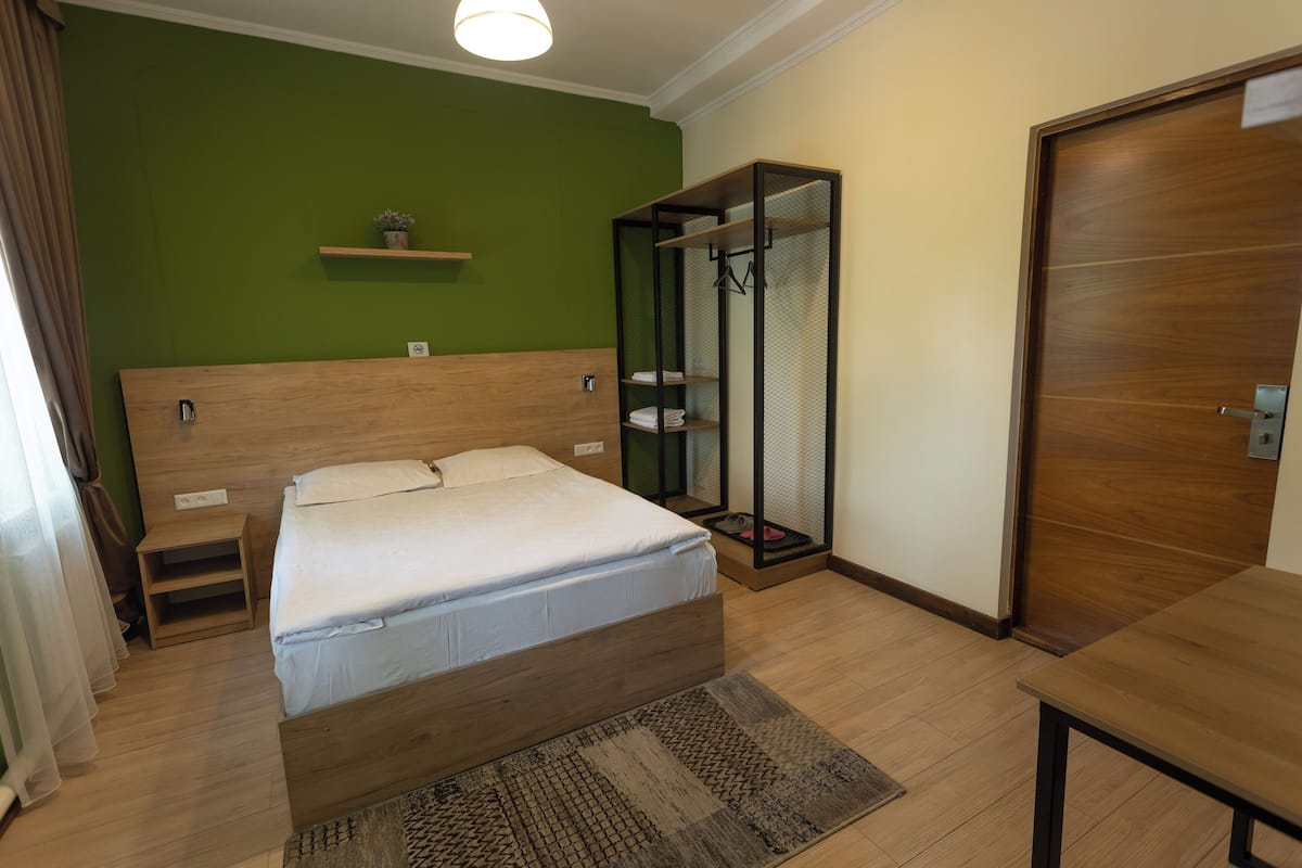 A bedroom with a wooden bed, a small nightstand, a shelf with a plant, a wardrobe, and a wooden table.