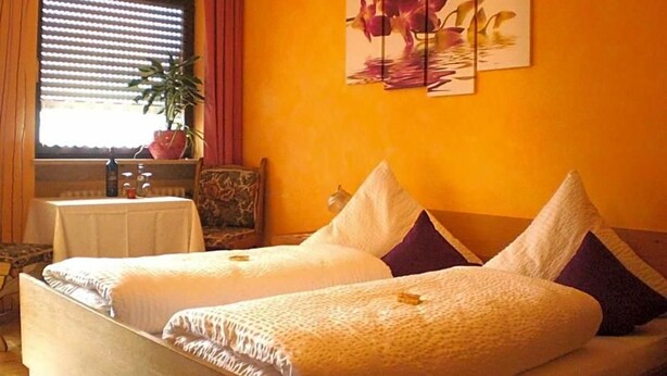 Ein Zimmer mit zwei Betten, einem Fenster mit Jalousien, einem Blumentopf und einem Bild an der Wand.