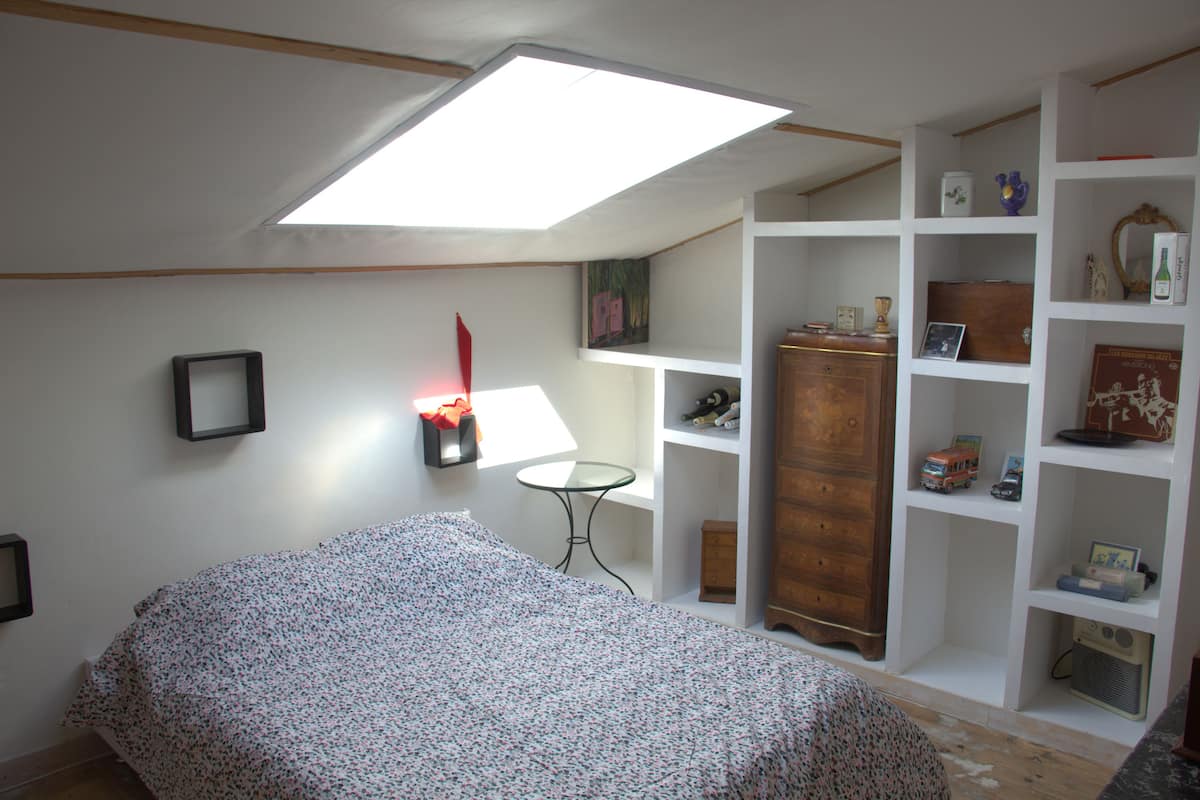 Un dormitorio con tragaluz, una cama, una mesita pequeña y un estante blanco con diversos objetos.