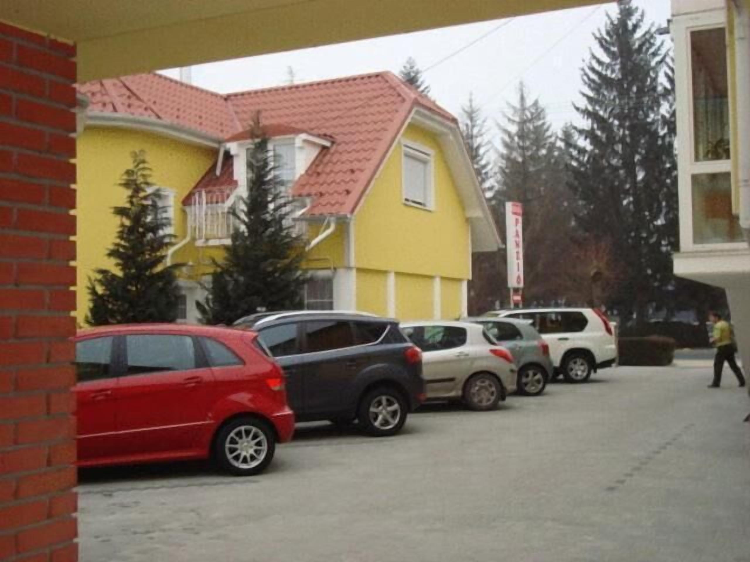 A parking area with several cars, a yellow building with a red roof, and a person walking.
