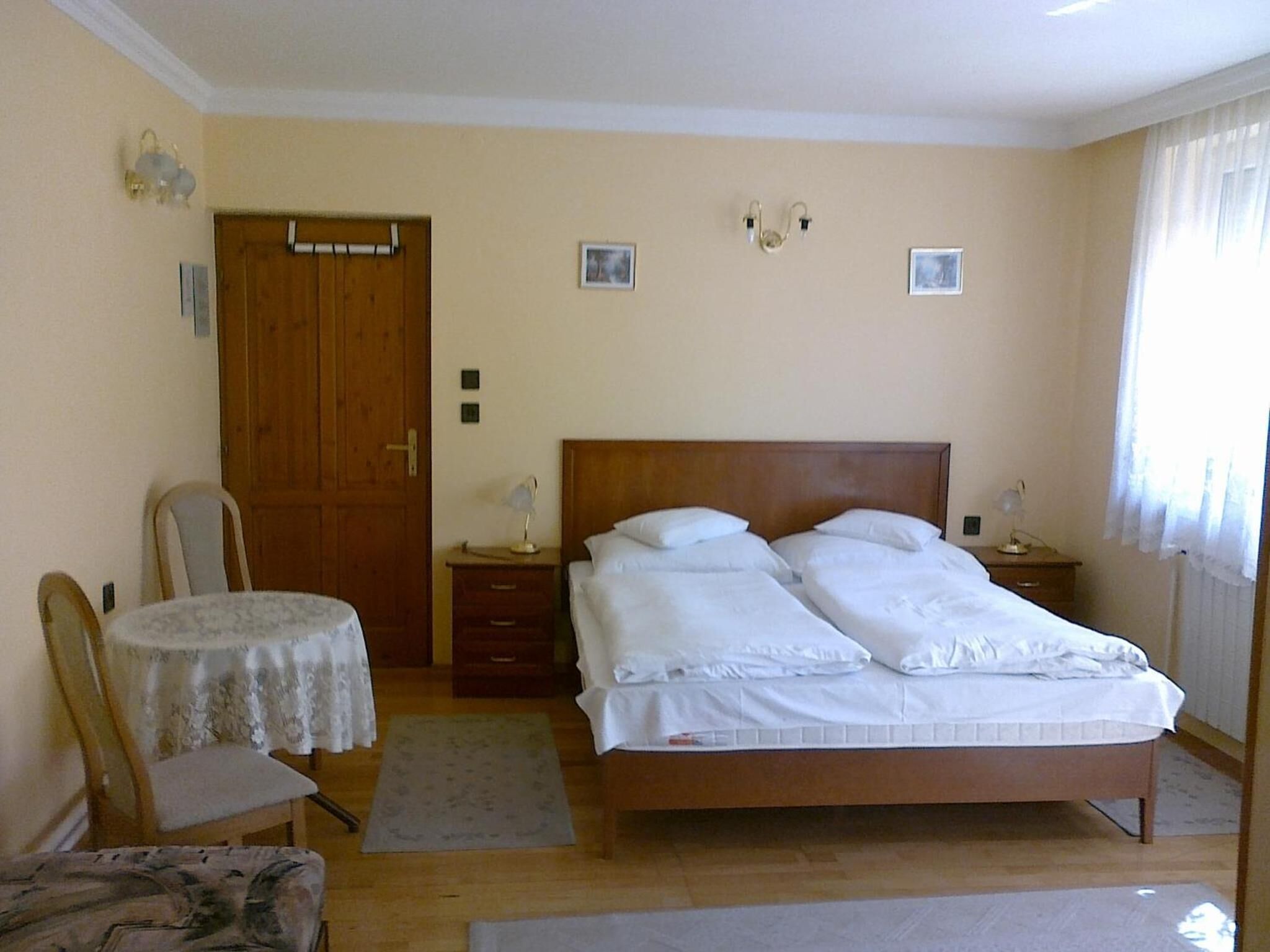 A bedroom with a double bed, a wooden door, a chair, a small table, and a nightstand.