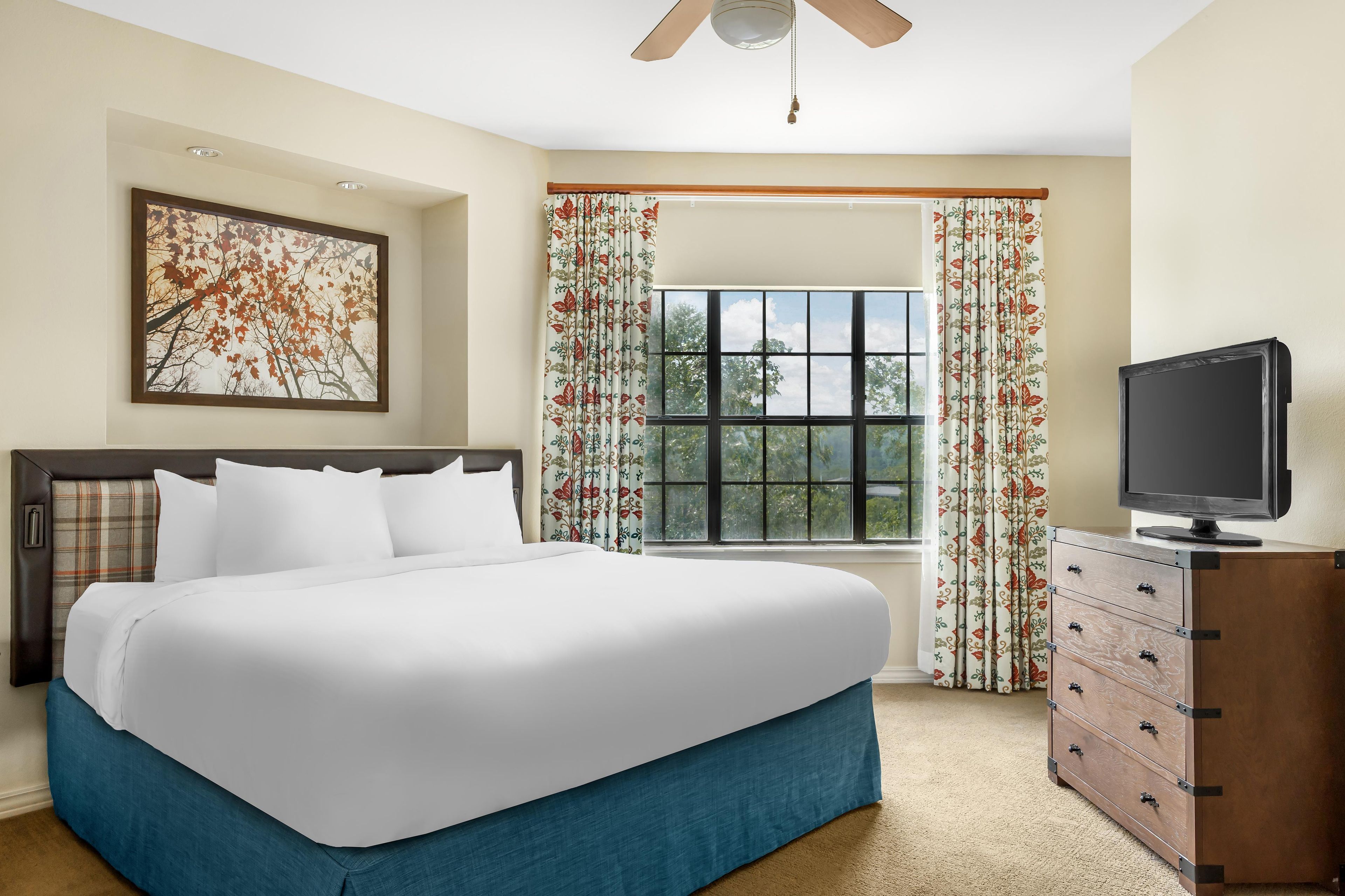 A hotel room with a large bed, a television on a wooden dresser, a window with floral curtains, and a ceiling fan.