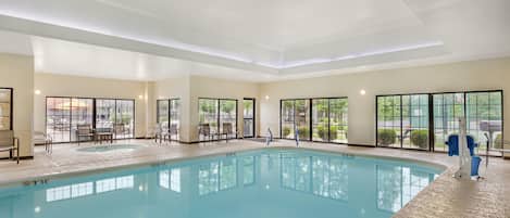 An indoor swimming pool with clear water, surrounded by lounge chairs and a view of the outdoors through large windows.