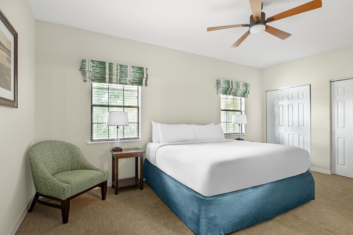 A hotel room with a bed, a chair, two windows with blinds, a ceiling fan, and a door.