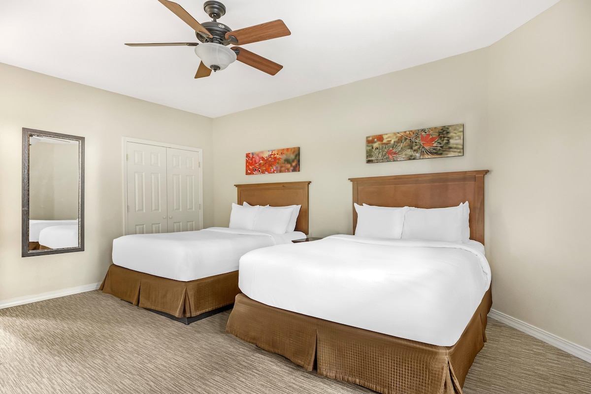A hotel room with two beds, a ceiling fan, a large mirror, and two wall-mounted paintings.