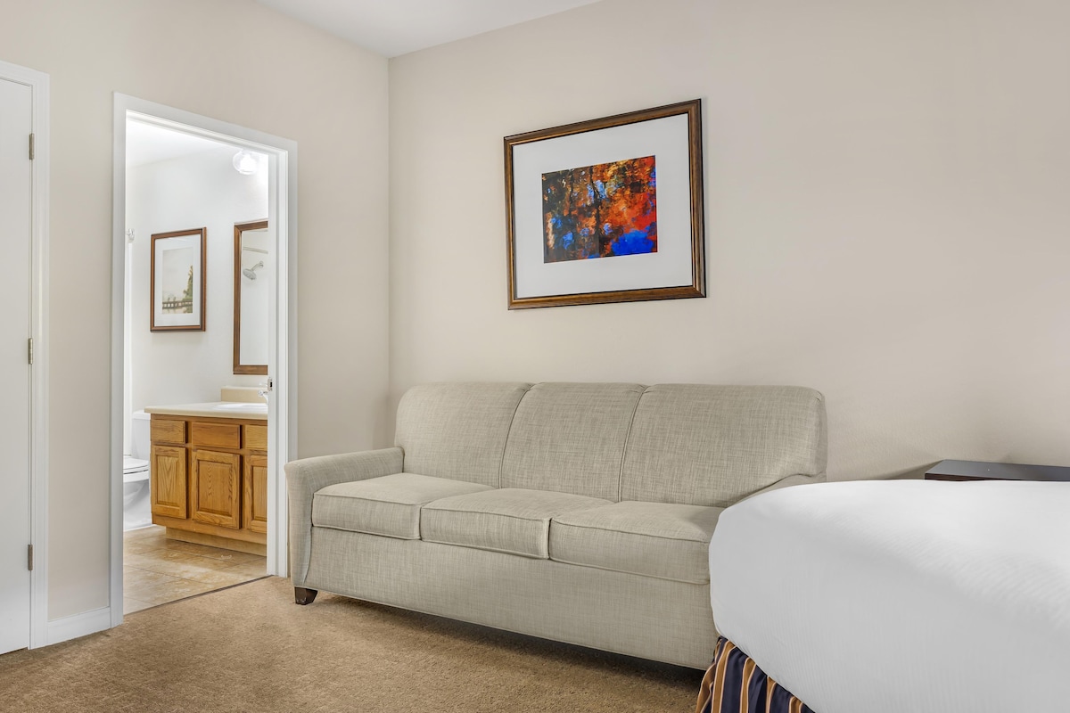 A hotel room with a beige sofa, a bed with white bedding, a wooden cabinet, and a framed abstract painting on the wall.