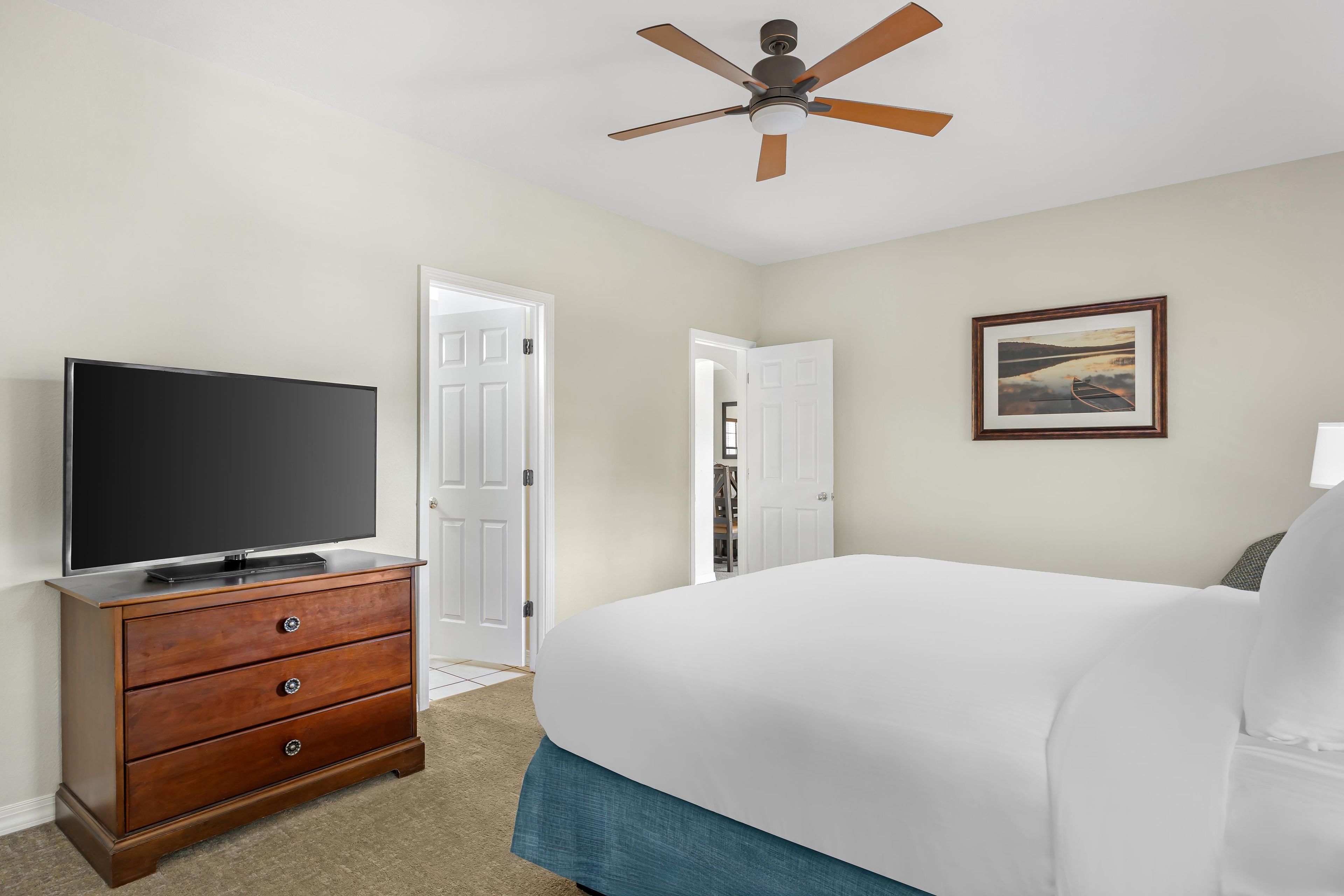 A hotel room with a flat-screen TV on a wooden dresser, a bed with white bedding, a ceiling fan, and a framed picture on the wall.