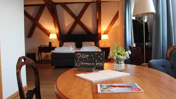 Une chambre d’hôtel avec un plafond en bois, un lit avec du linge de lit blanc, une table de chevet avec une lampe, une petite table avec un vase de fleurs et une chaise.