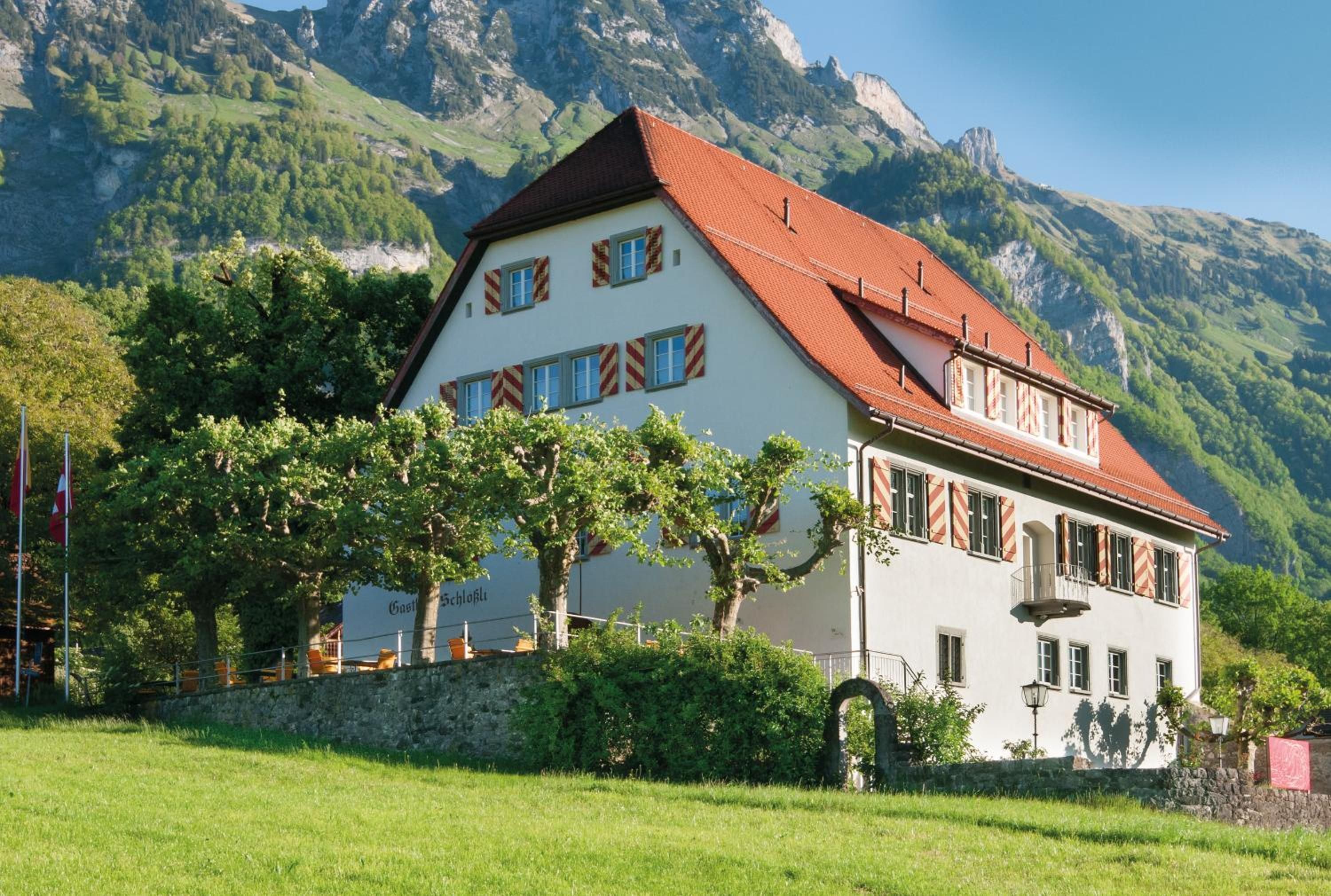 A two-story building with a red-tiled roof, surrounded by greenery and mountains.
