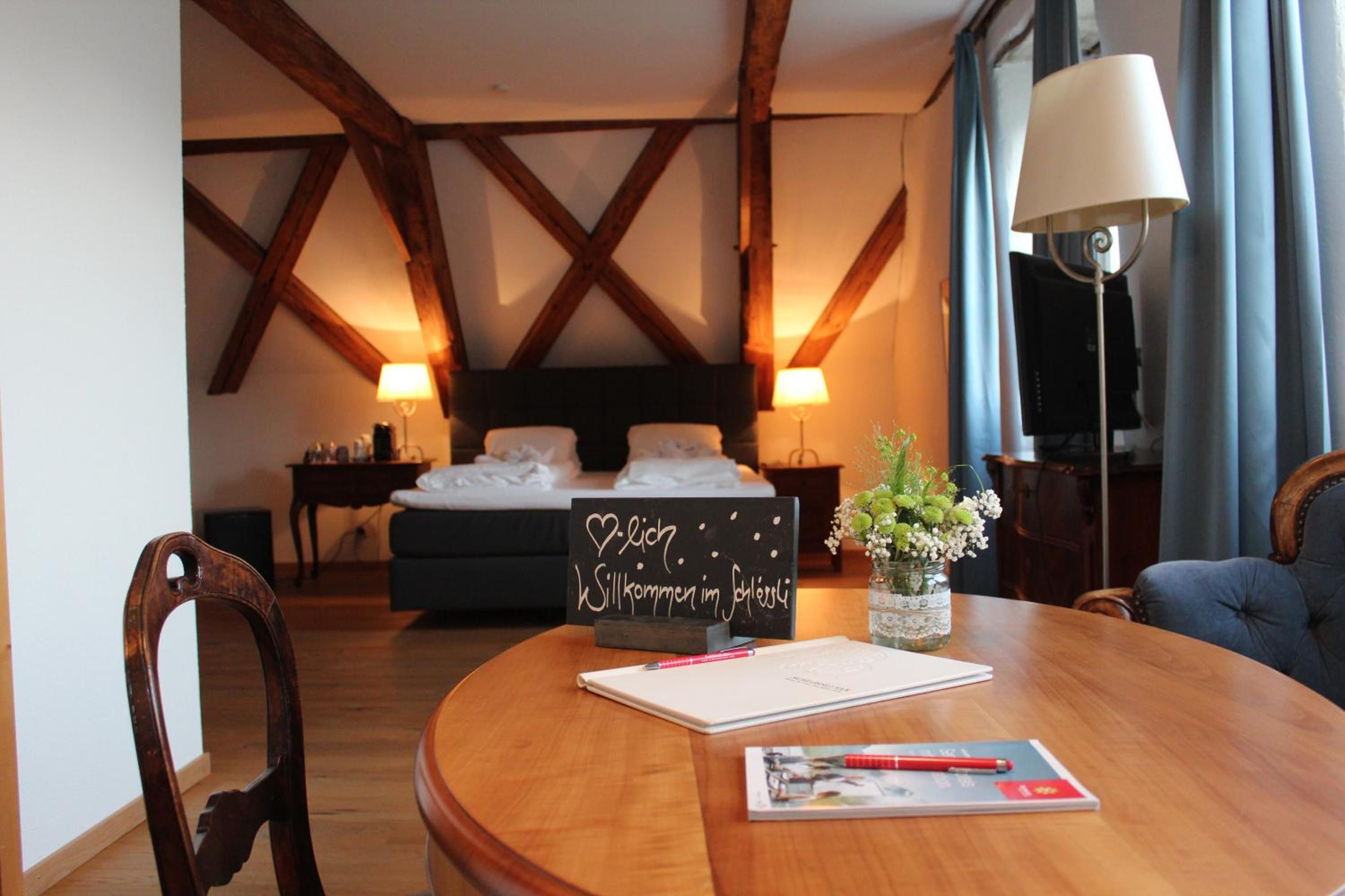 A hotel room with a wooden ceiling, a bed with white linens, a bedside table with a lamp, a small table with a vase of flowers, and a chair.