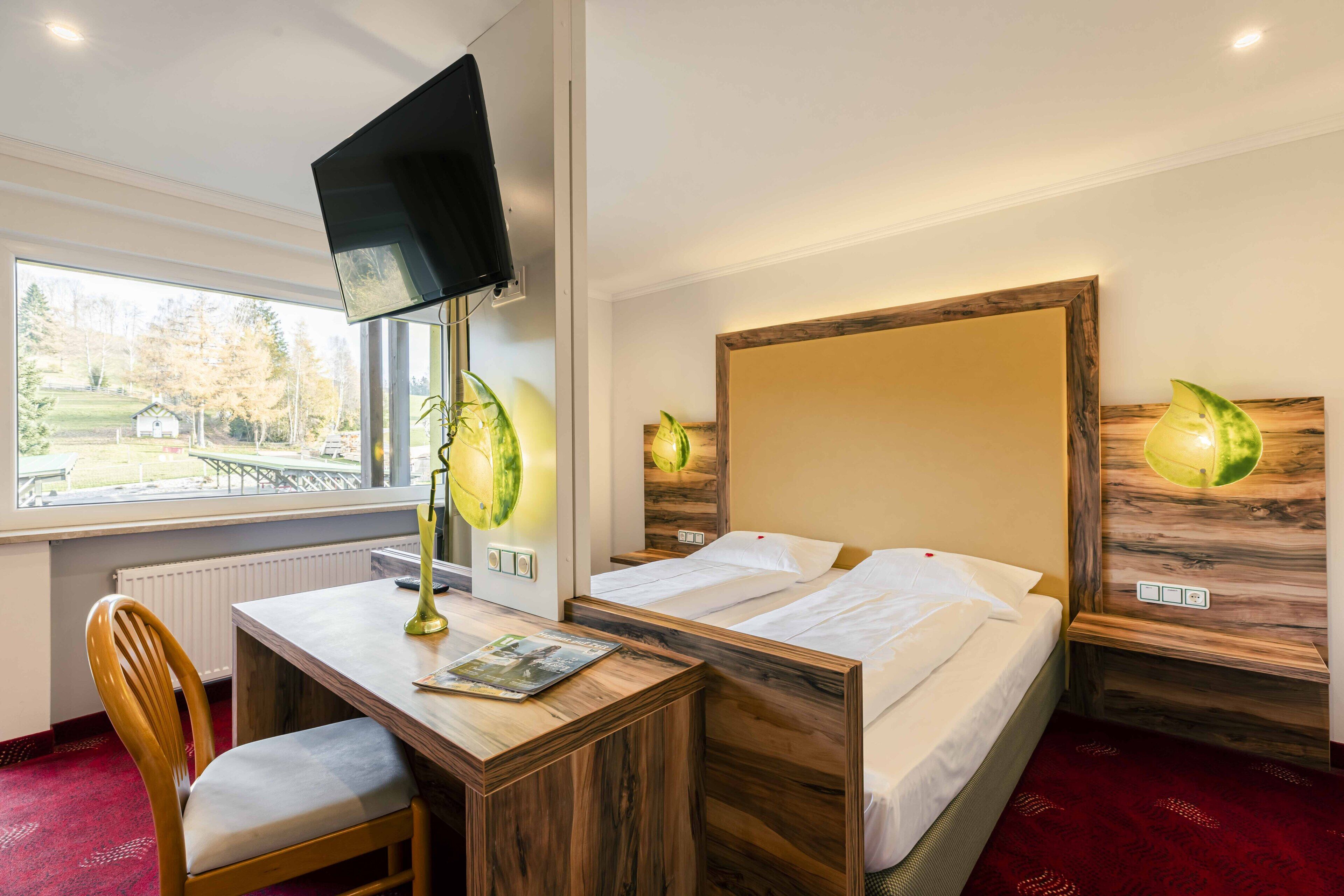 Une chambre d’hôtel avec un lit en bois, un bureau avec une chaise, une télévision et une fenêtre donnant sur un parc.