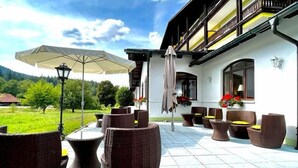 An outdoor patio with wicker furniture and umbrellas, a balcony with a yellow railing, and a view of a green landscape. - Garni GrünWies - Adults Only (Lohberg)