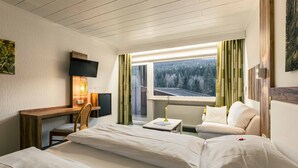 A hotel room with a bed, a desk with a TV, a chair, a small table with a lamp, and a view of a forest through the window. - Garni GrünWies - Adults Only (Lohberg)