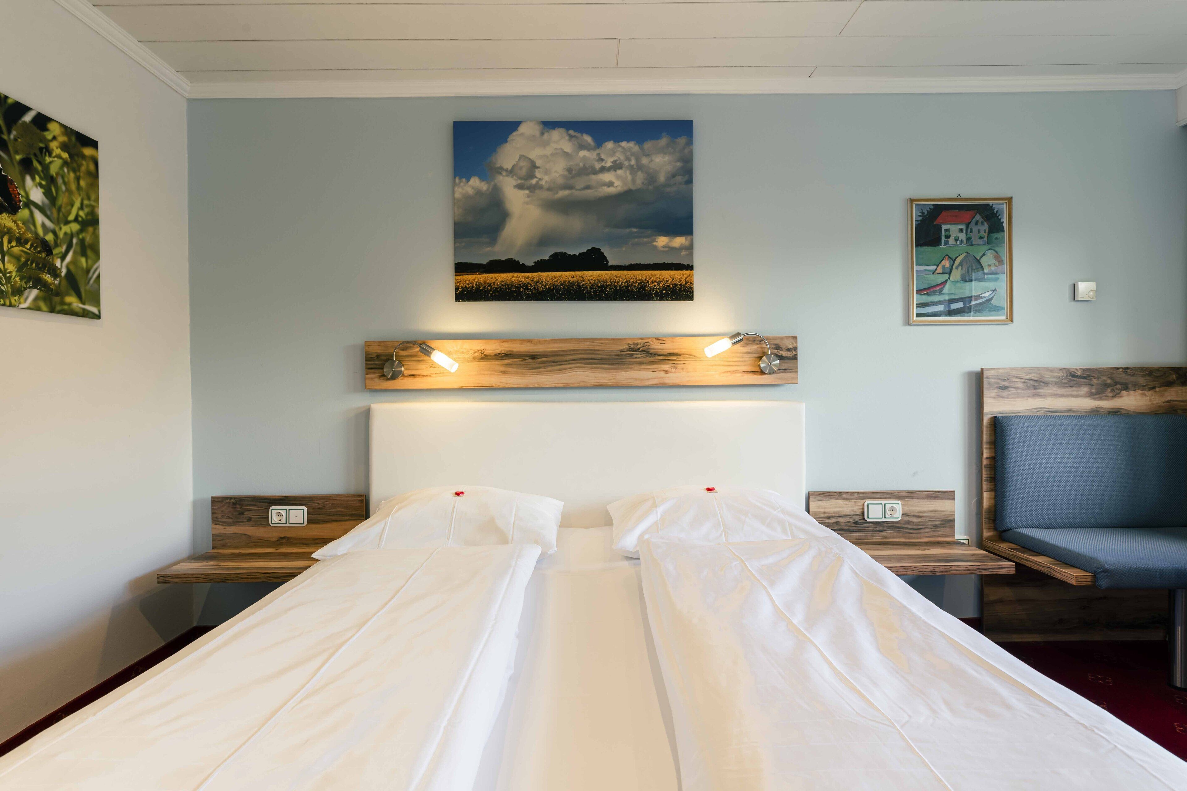 A double bed with white linens, a wooden headboard, and a mounted painting of a landscape.