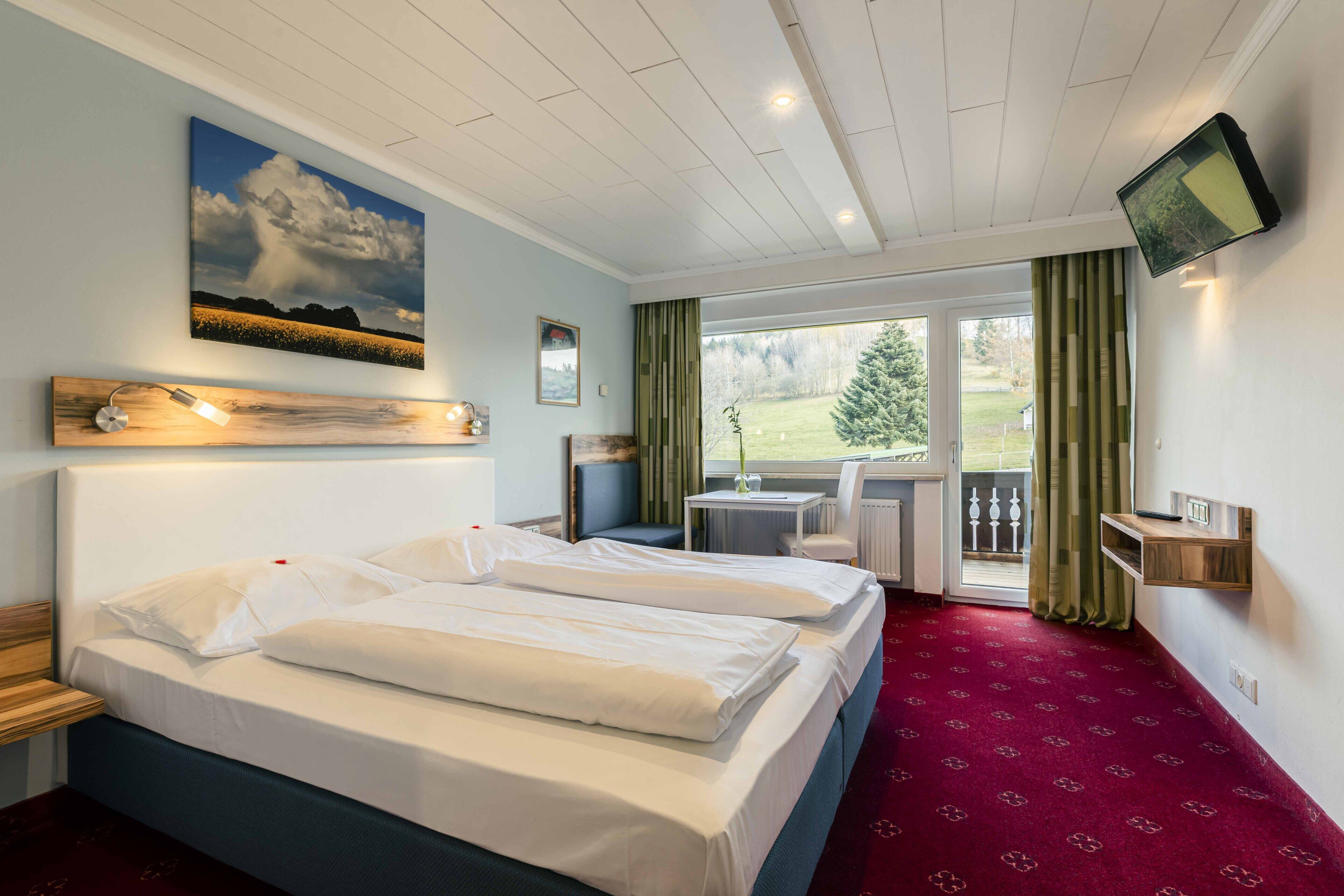 Ein Hotelzimmer mit zwei Betten, einem Fernseher, einem Fenster mit Vorhängen und Blick auf eine grüne Landschaft.
