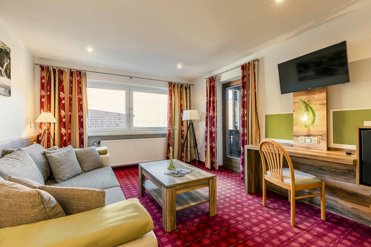 Une chambre d’hôtel avec un canapé, une table basse en bois, une chaise, un téléviseur et une fenêtre avec des rideaux.