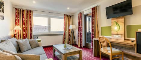 Habitación de hotel con sofá, una mesa de centro de madera, una silla, televisor y ventana con cortinas.