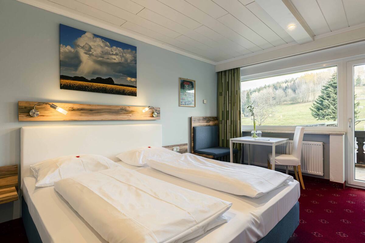 Une chambre d’hôtel avec un lit, un bureau, une chaise, une fenêtre donnant sur une forêt et un tableau accroché au mur.