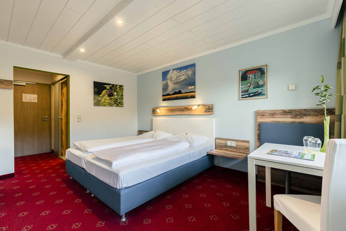 Une chambre d’hôtel avec un lit simple, une petite table avec une chaise, une tête de lit en bois et un sol recouvert d’une moquette rouge.