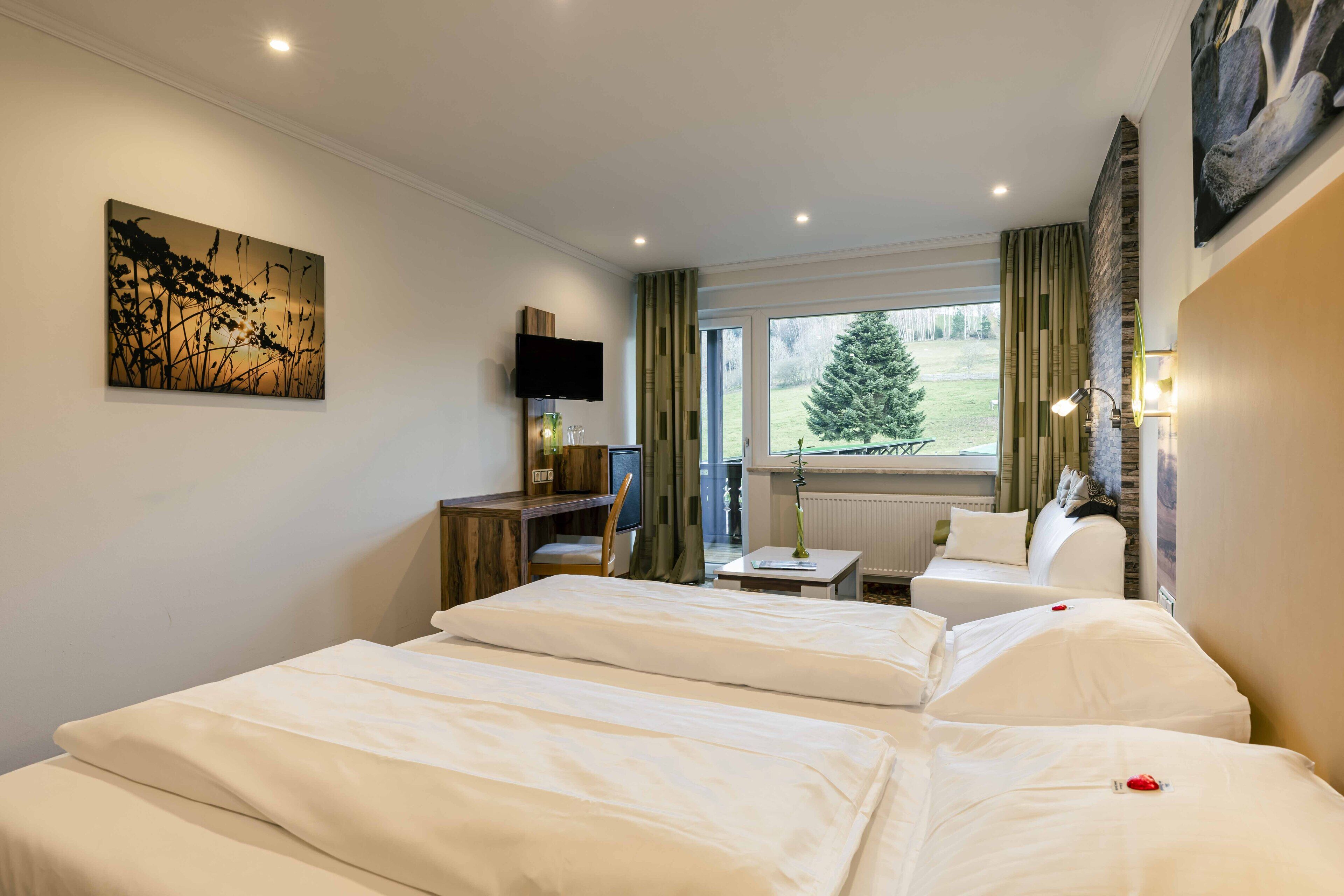 Ein Hotelzimmer mit zwei Betten, einem Schreibtisch, einem Fernseher und einem Fenster mit Blick auf einen Baum.