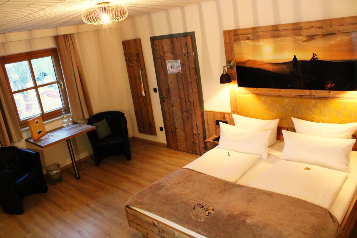 Une chambre à coucher avec une tête de lit en bois, un lit recouvert de draps blancs, un bureau avec une chaise et une fenêtre avec des rideaux.