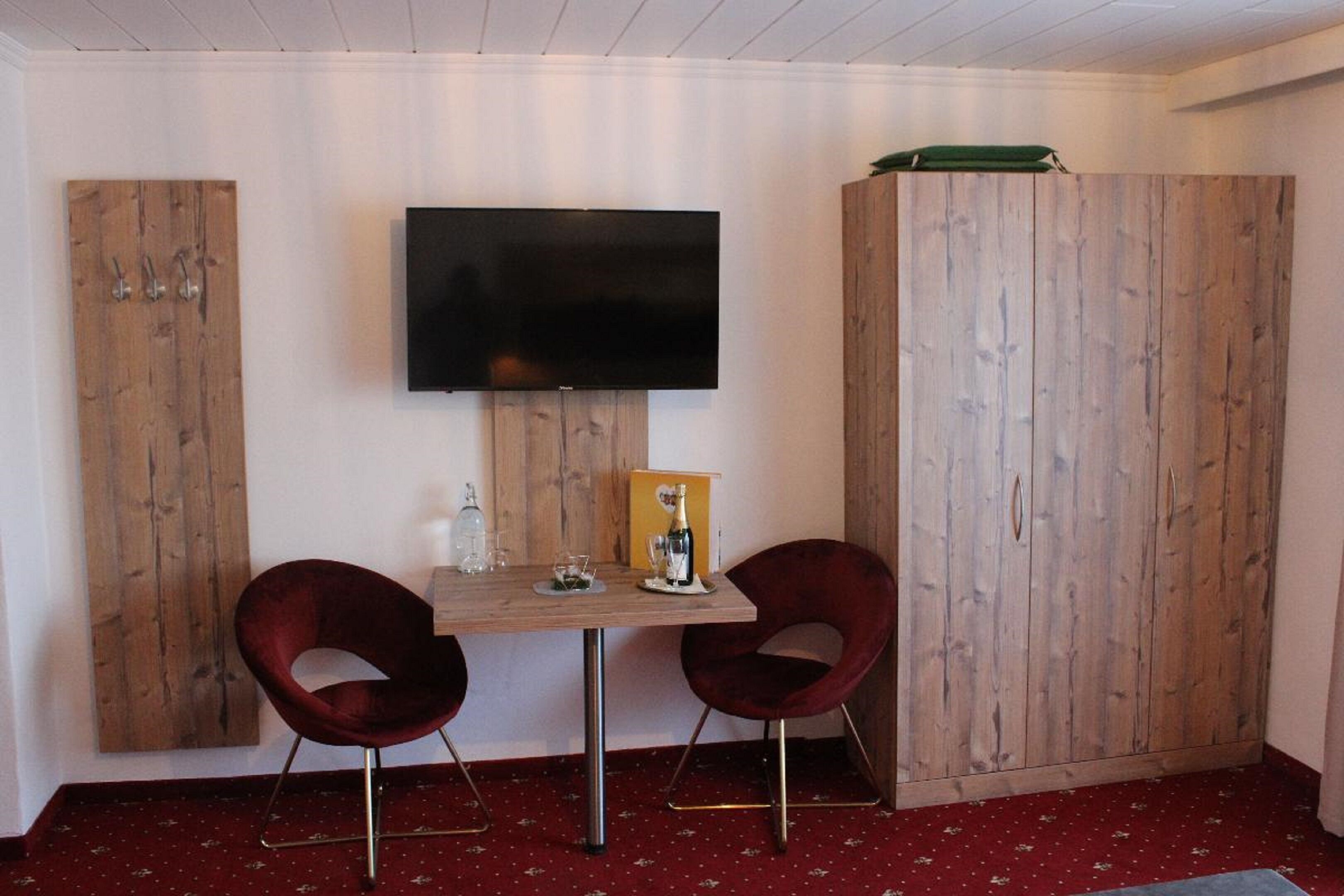 A room with a wooden table, two red chairs, a flat-screen TV mounted on the wall, and wooden wardrobes.