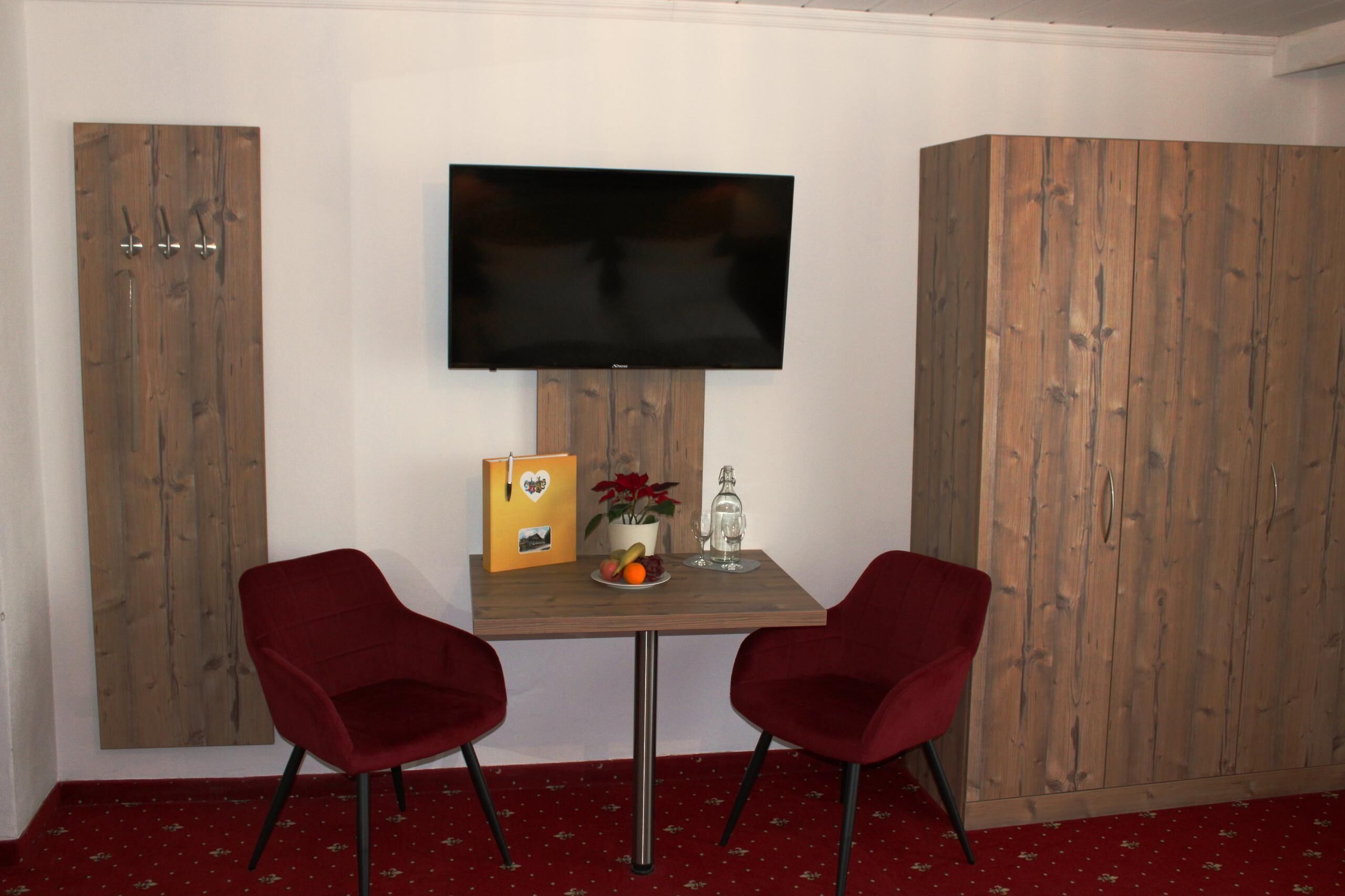 A room with a wooden wardrobe, a television mounted on the wall, a small table with two red chairs, and a red carpet.