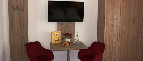A room with a wooden wardrobe, a television mounted on the wall, a small table with two red chairs, and a red carpet.