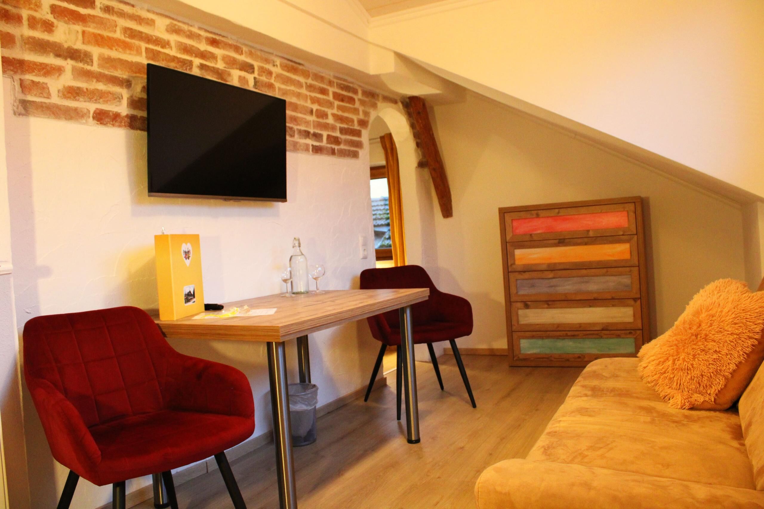 A cozy room with a brick wall, a flat-screen TV, a wooden table, red chairs, and a sofa with orange cushions.