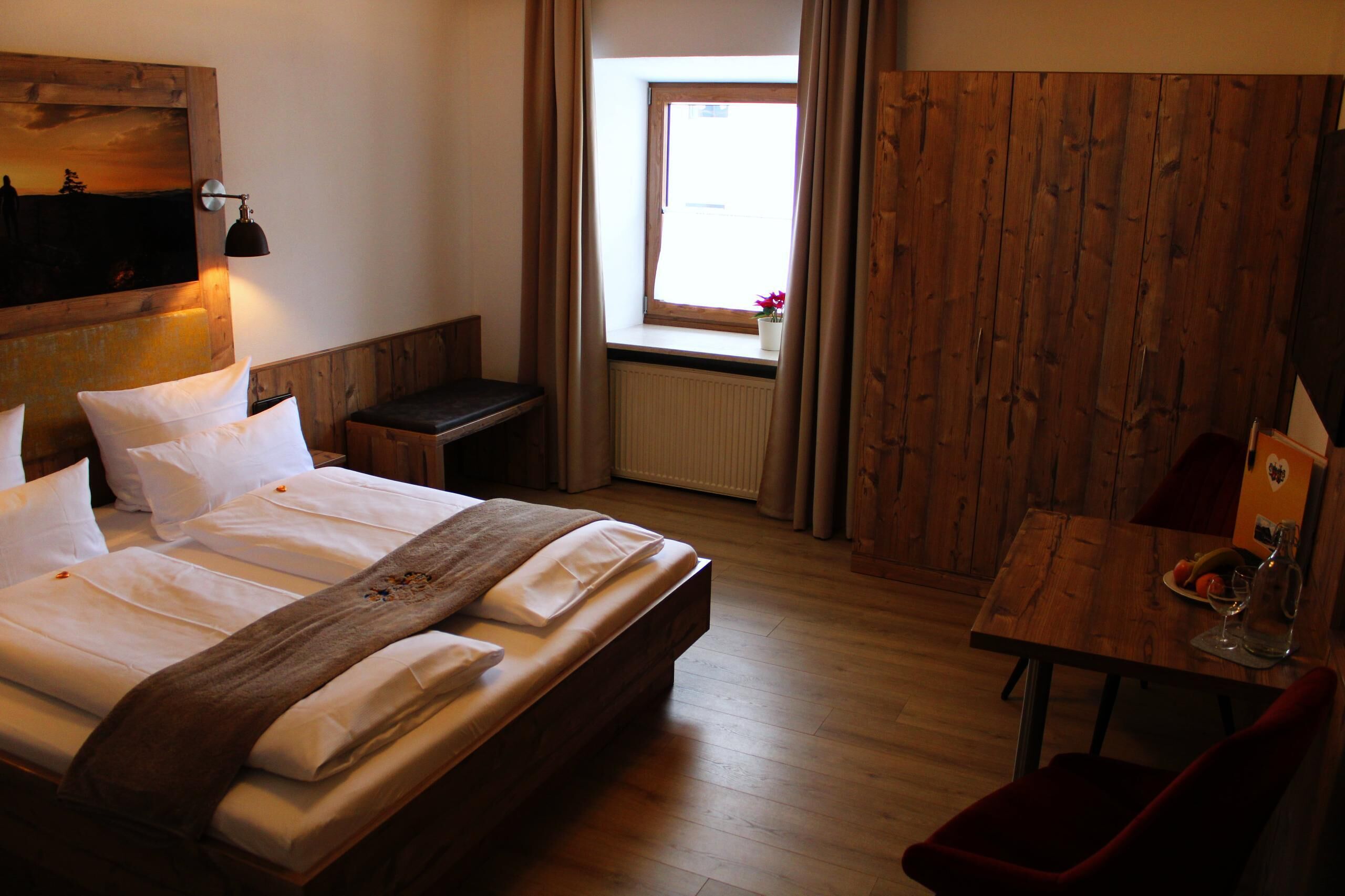 Una habitación de hotel con una cama de madera, una mesita con una botella y un vaso, una ventana con cortinas y una pared de madera.