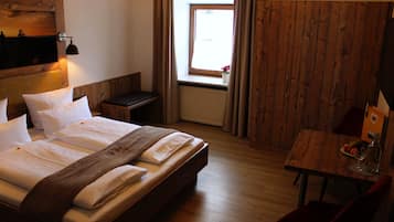 A hotel room with a wooden bed, a small table with a bottle and glass, a window with curtains, and a wooden wall.