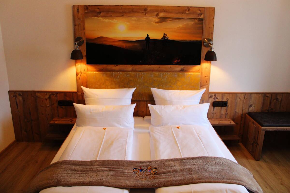 Un lit double avec du linge de lit blanc, deux tables de chevet, une tête de lit en bois ornée d’une peinture représentant un coucher de soleil, et deux lampes fixées au mur.