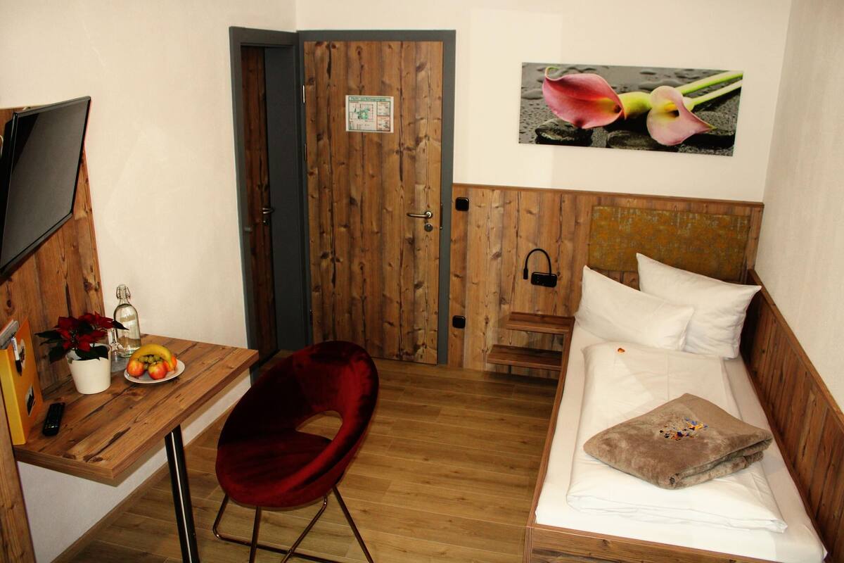 Une pièce de taille modeste, avec un bureau en bois, une chaise rouge, un lit avec une literie blanche et un téléviseur fixé au mur.