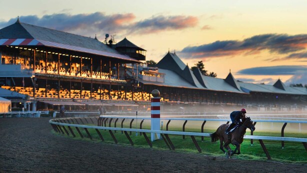 Un hippodrome avec un cavalier à cheval, entouré de tribunes et d’un grand bâtiment doté d’un toit remarquable.