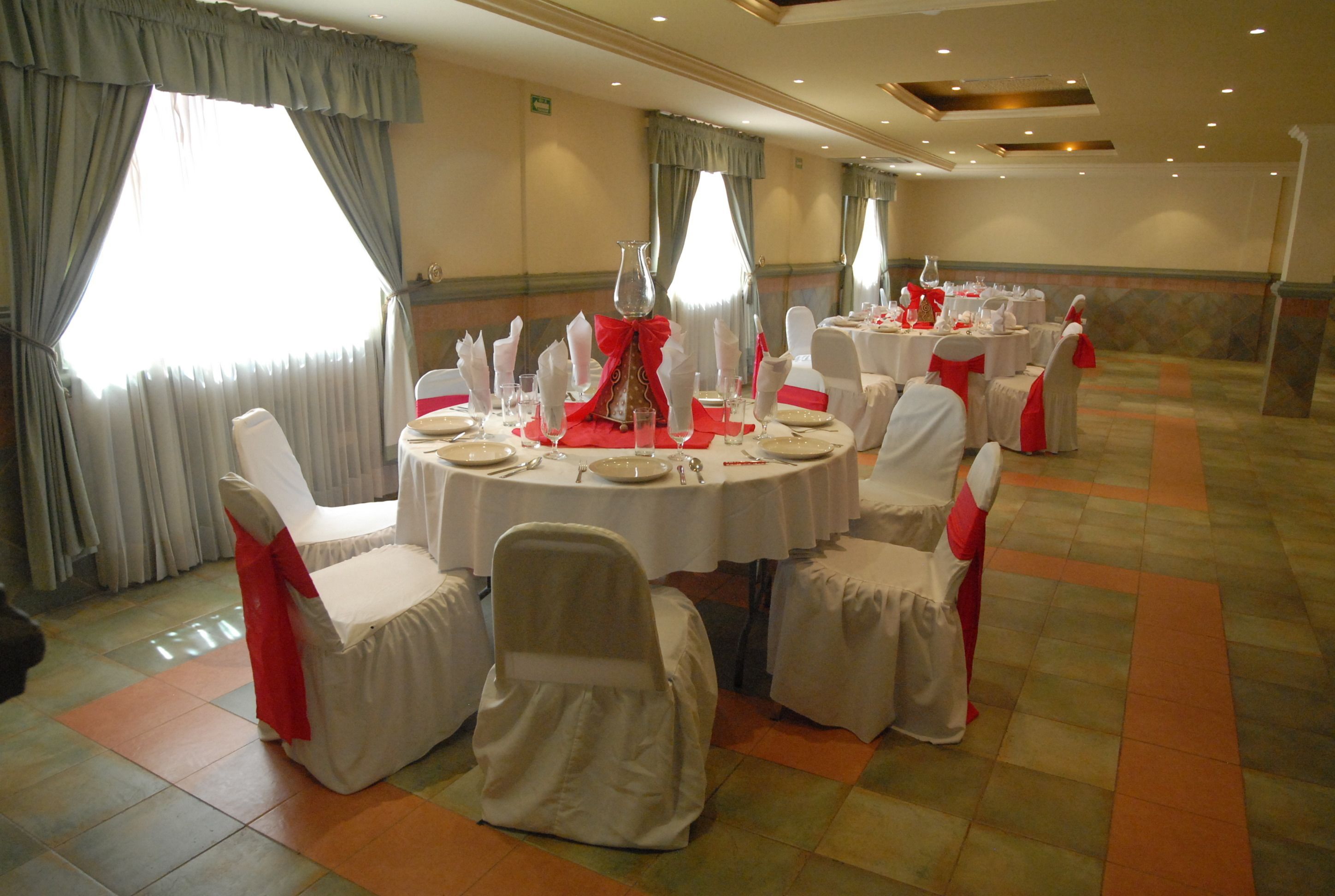 Un salón de banquetes con mesas redondas dispuestas para comer, sillas con fundas blancas y cintas rojas, y ventanas amplias con cortinas.