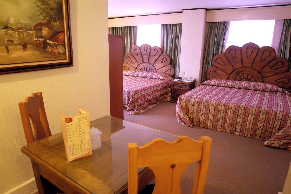 A hotel room with two beds, a dining table, and a chair. There is a painting on the wall and a window with curtains.