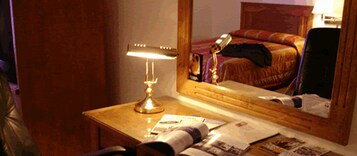 Una habitación de hotel con un escritorio de madera, una lámpara, un espejo que refleja una cama con una manta roja y algunos papeles sobre el escritorio.