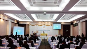 A conference room with rows of tables and chairs, a stage with a podium, and a large screen displaying a presentation. - City Park Hotel (Nakhon Ratchasima)