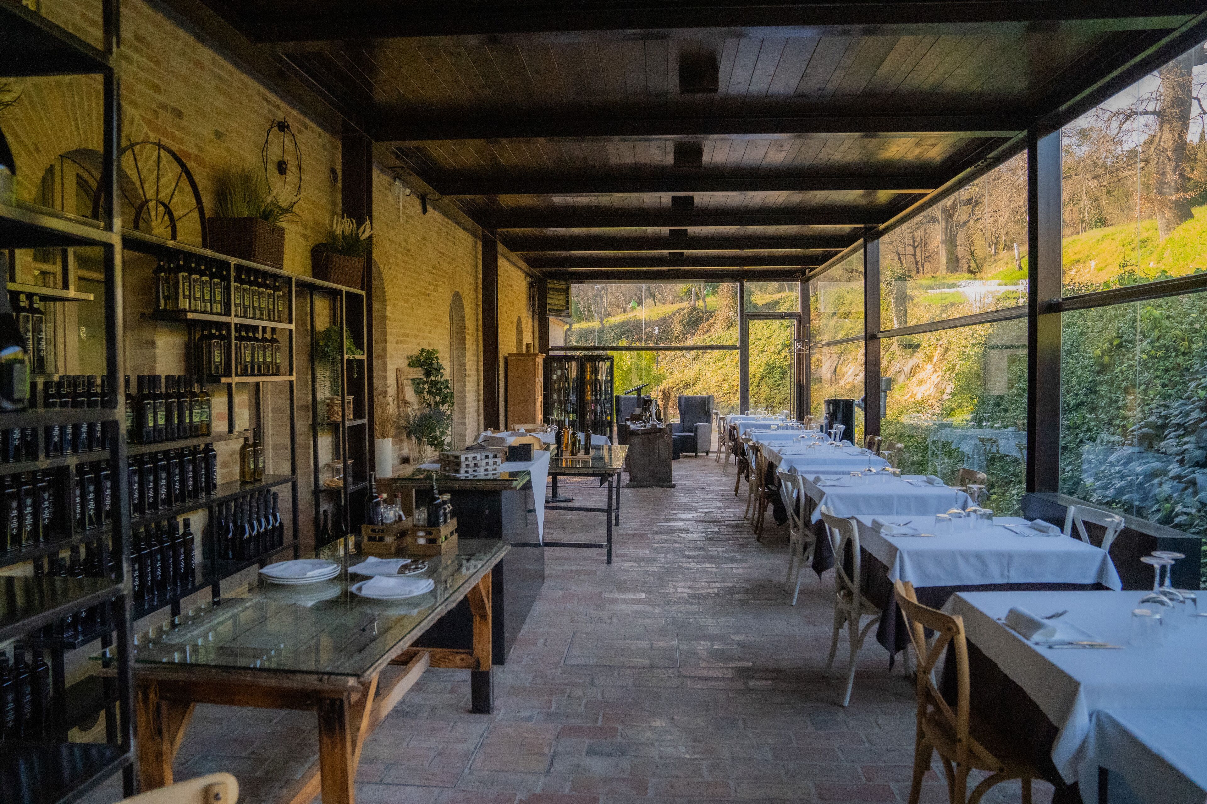 Un espace extérieur abrité, aménagé pour les repas, avec des tables et des chaises, un mur de briques orné d’étagères remplies de bouteilles, et une vue sur la verdure environnante.