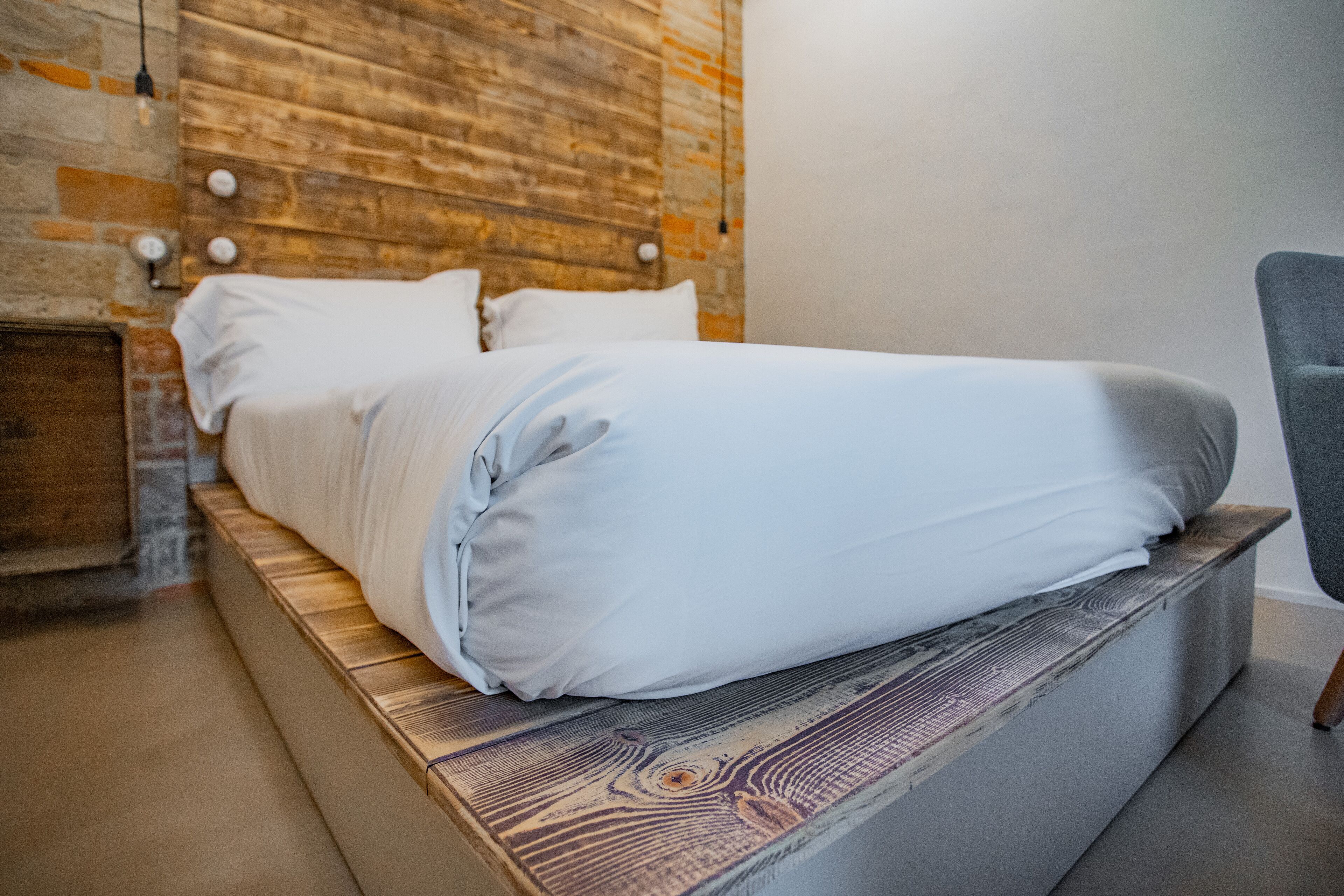 Un lit avec une tête de lit en bois et une literie blanche, dans une pièce dont un mur est en briques et qui est éclairée par un luminaire moderne.