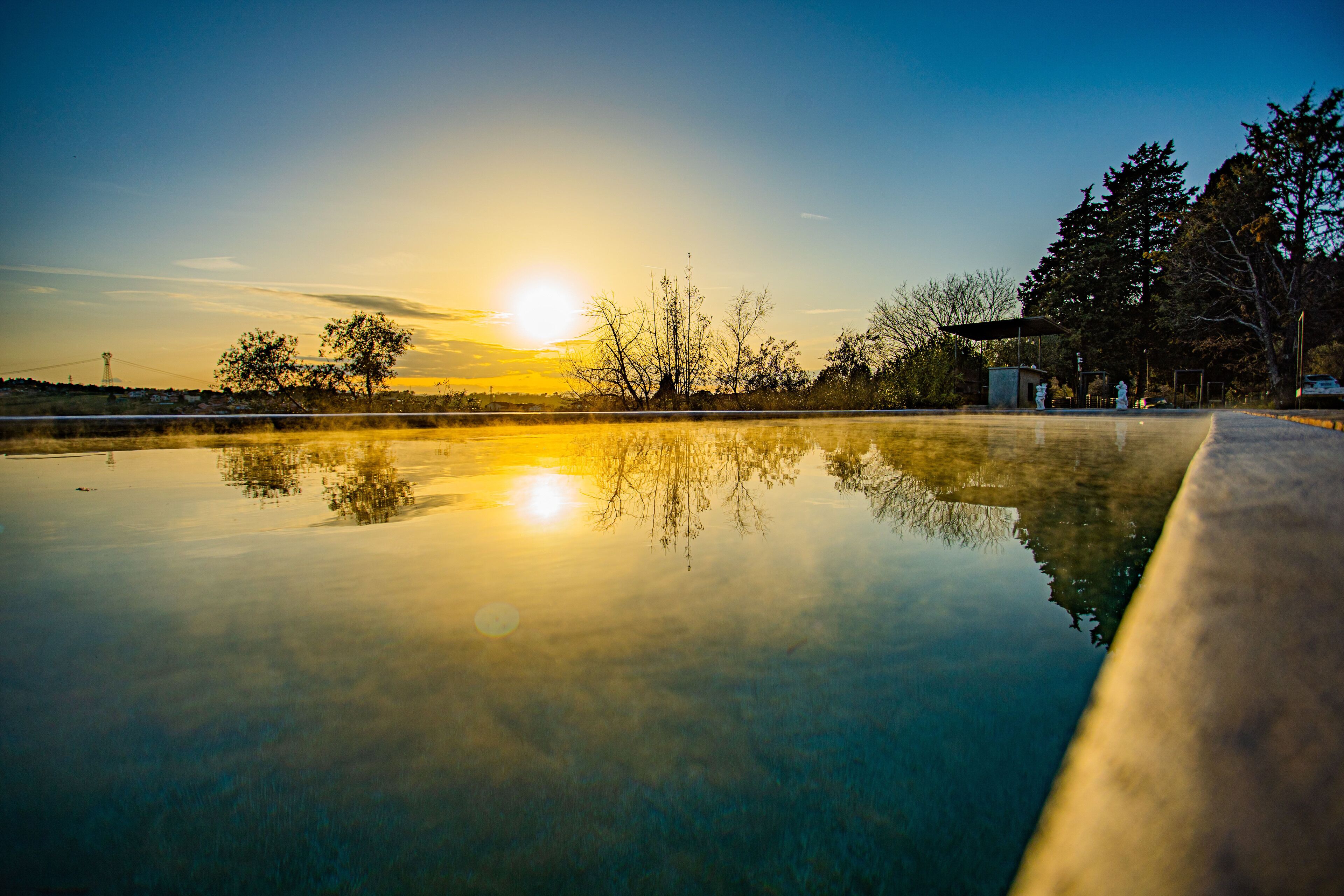 A serene outdoor pool area with a clear reflection of the sunset, surrounded by trees and a distant power line structure.