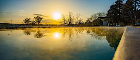 A serene outdoor pool area with a clear reflection of the sunset, surrounded by trees and a distant power line structure.