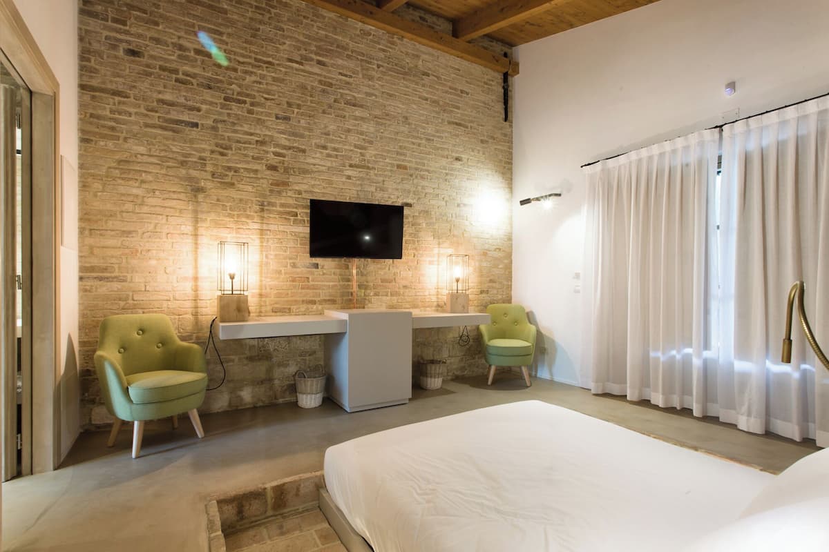 A hotel room with a brick wall, a flat-screen TV, a bed, two chairs, and a window with curtains.