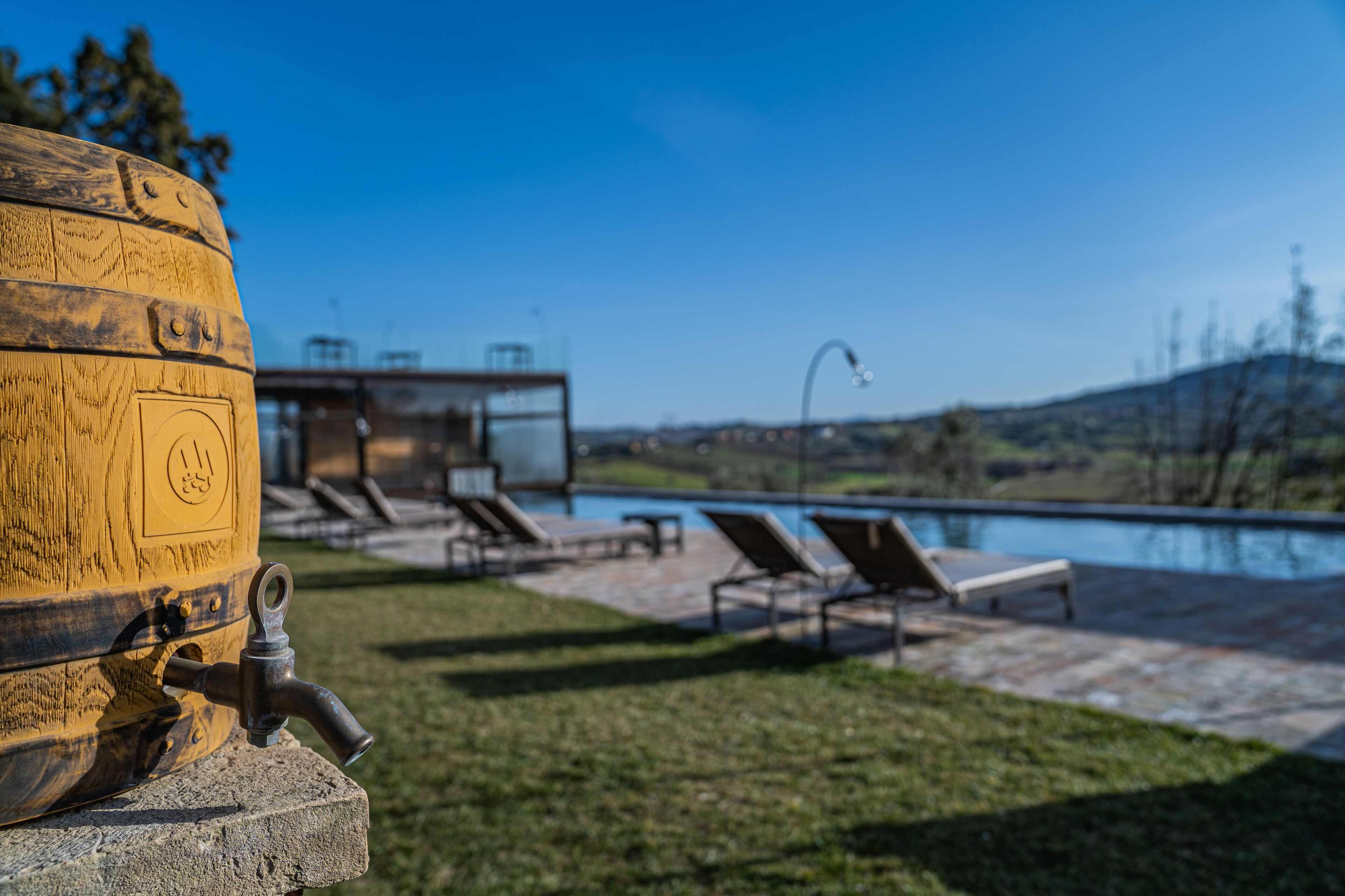 A wooden barrel with a tap, a pool, and lounge chairs in a sunny outdoor setting.