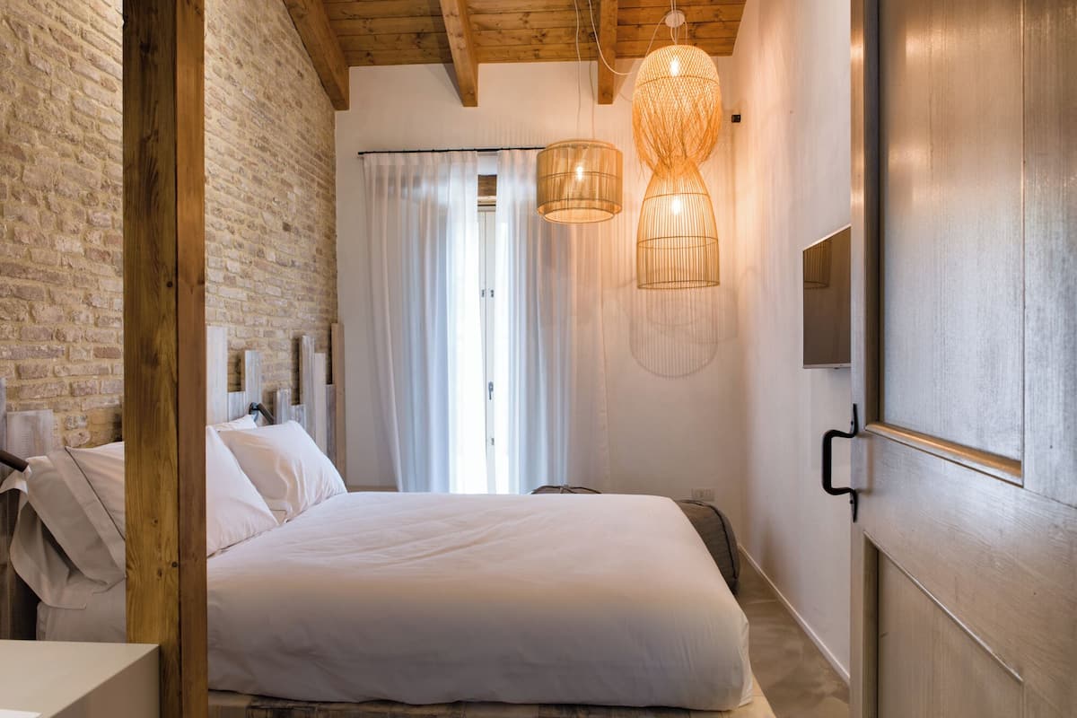 A bedroom with a large bed, wooden beams, a brick wall, and two hanging light fixtures.