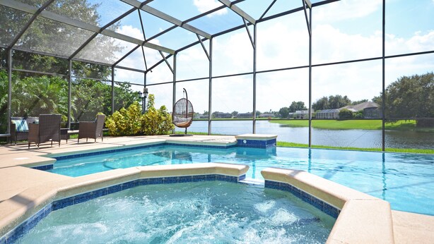 A swimming pool with a glass enclosure, surrounded by lounge chairs and a hanging chair, with a view of a lake and greenery.