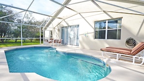 En swimmingpool med buet kant, omgivet af en overdækket patio med liggestole og et bord.