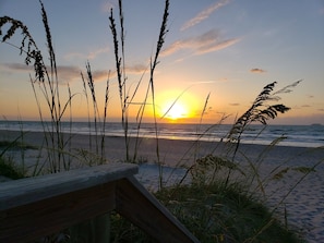 Una terraza de madera con vista a una playa, con hierbas altas al frente y un atardecer sobre el océano.